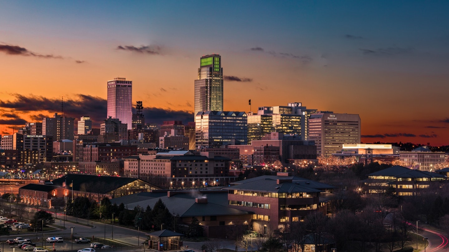 The skyline of Omaha Nebraska is seen at sunset with a striking orange and blue sky and lights glittering in the city