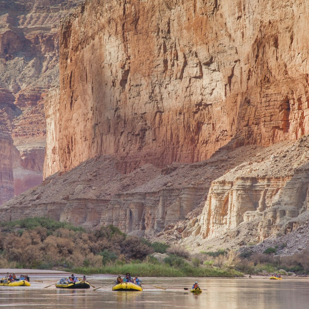 Whitewater rafts and a kayak drift on a calm part of the Colorado River in the Grand Canyon, as sheer rock walls fill the frame behind.