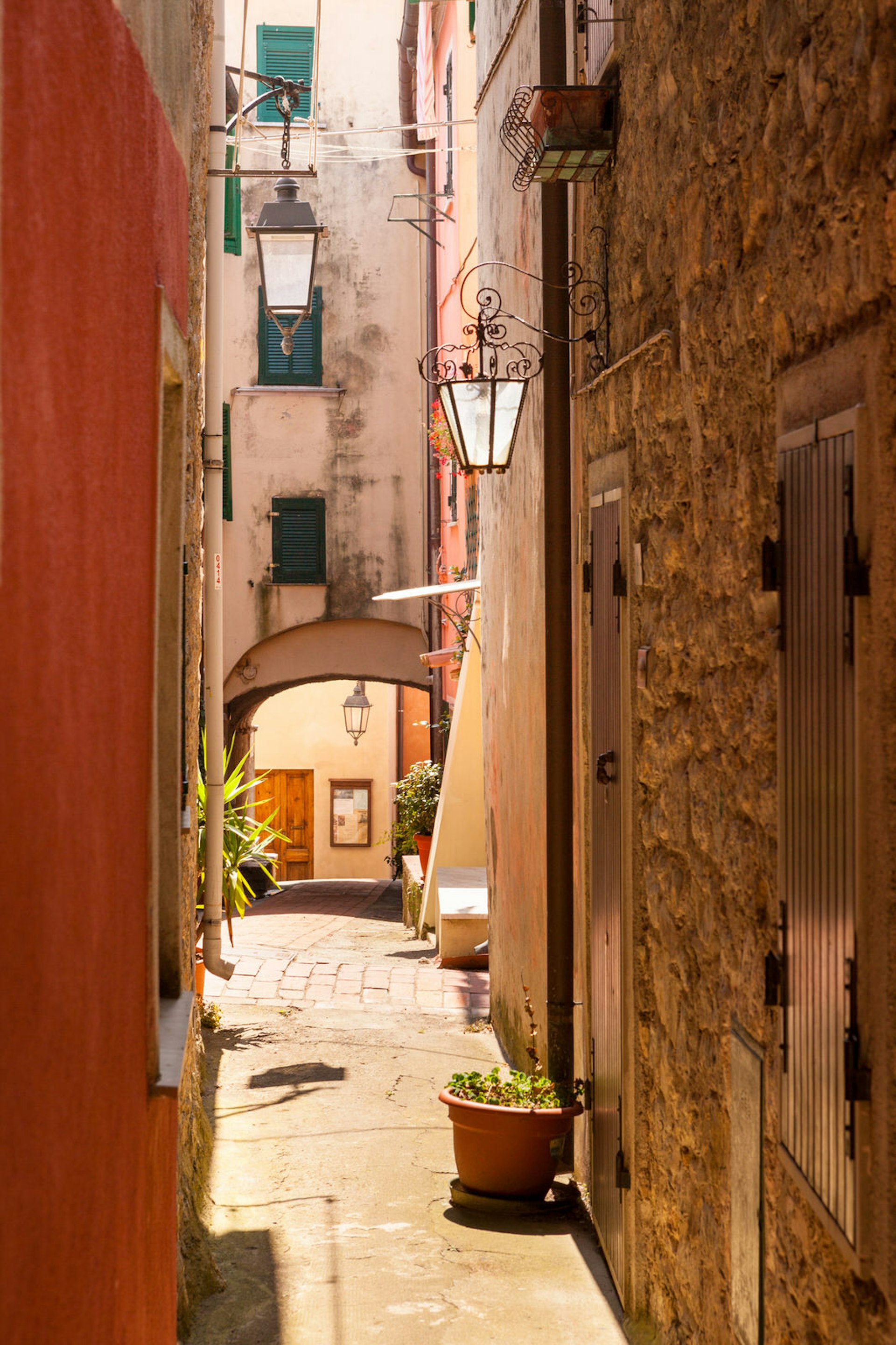 Looking down a narrow alleyway in Montemarcello, with glass lanterns and green shutters. There are some potted plants on the paved alley. The wall on the left is smooth and painted orange, the wall on the right is made from exposed, beige stones. 