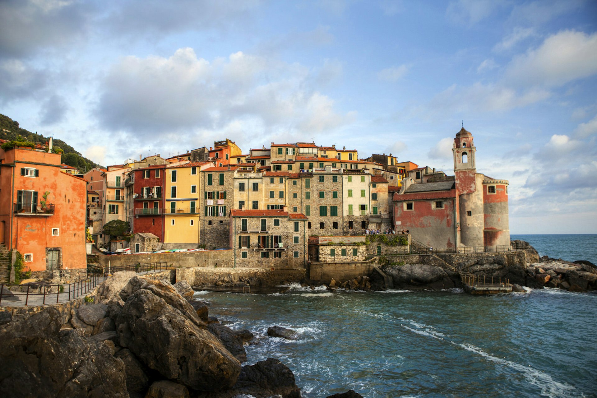 Tellaro's small harbour on a slightly overcast day. Narrow brick buildings are crammed in, side by side on a rocky outcrop. The buildings are warm yellows, beige, orange and terracotta. Closest to the sea there is a faded pink church. The sea is restless.