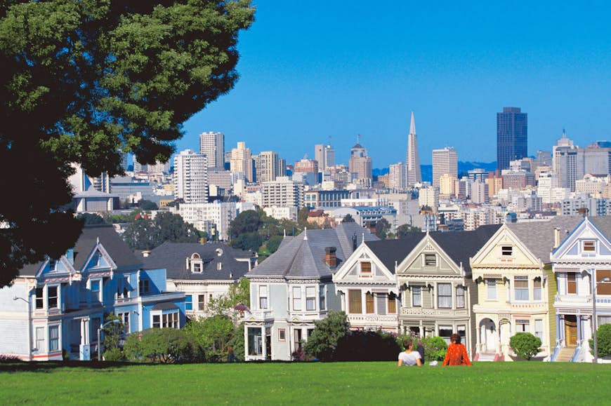 A man and woman lay in green grass looking at Victorian houses with the San Francisco skyline in the background