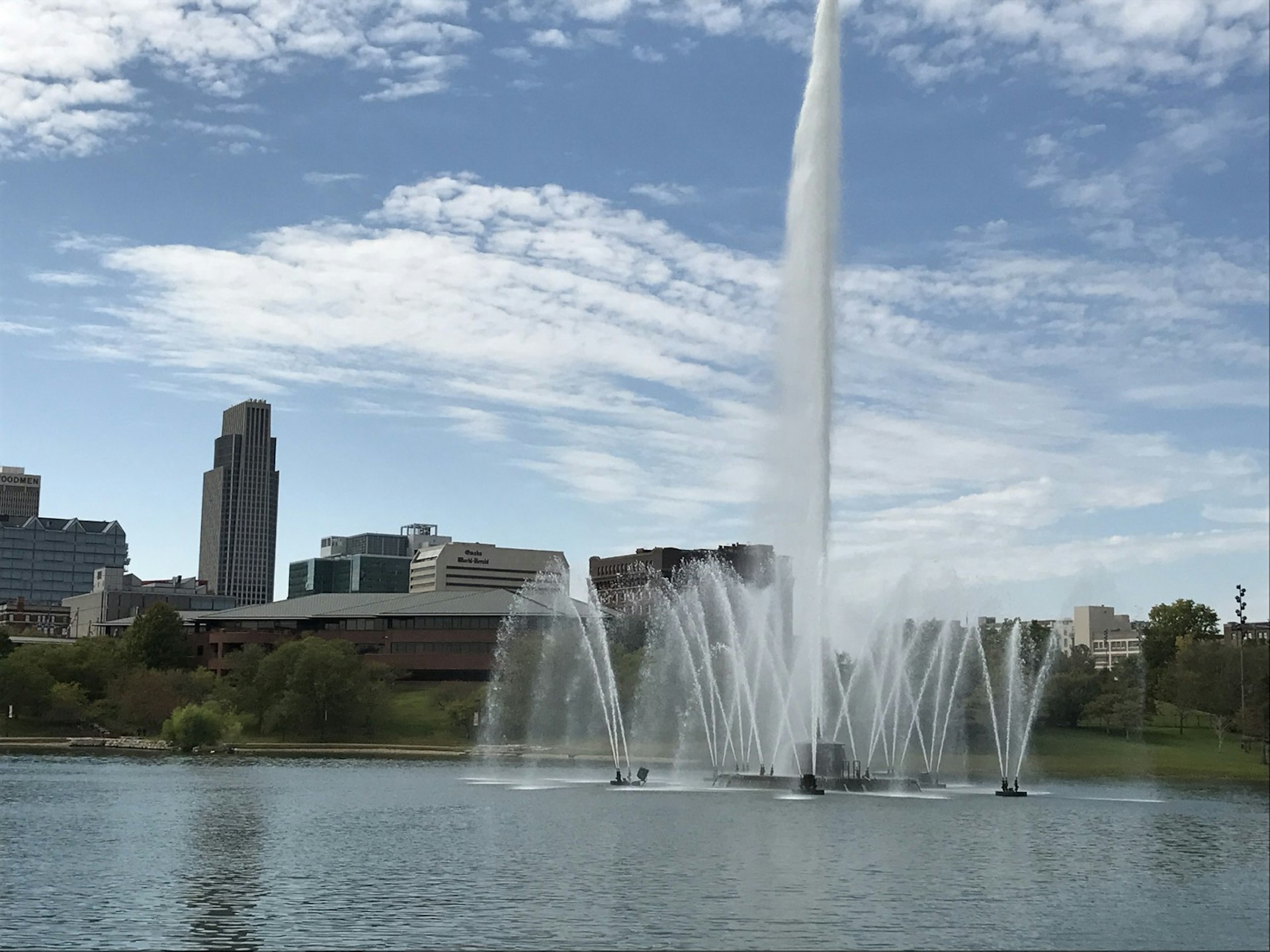 A large fountain dances in the middle of a lake with the Omaha skyline rising in the background
