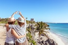 two women with their arms around each other create a heart with their hands while looking at a Mayan ruin on the beach