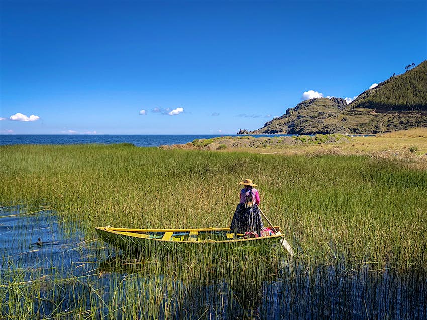 A woman in traditional Bolivian dress guides a yellow and green boat through a reed marsh on a sunny day in Isla Pariti, Bolivia