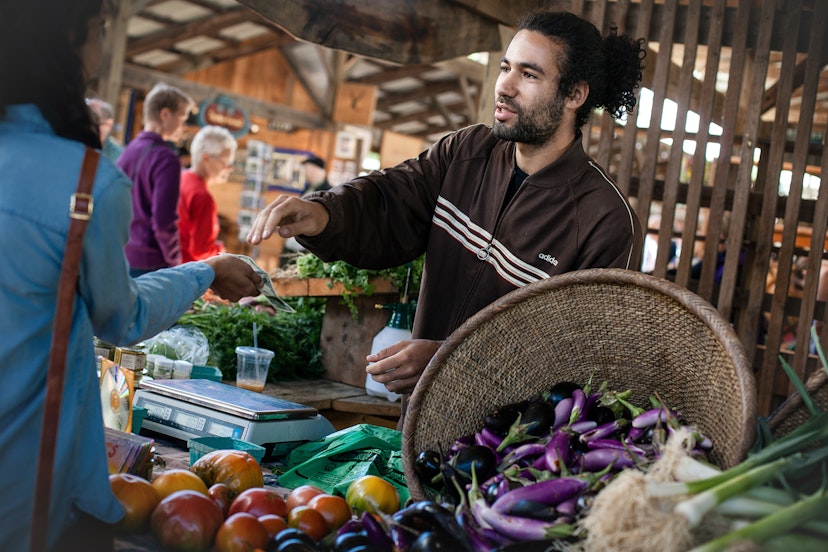 A man smiles and hands change to a woman over a stall of fresh local vegetables (eggplant, etc) at a farmer's market