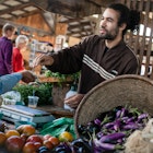 A man smiles and hands change to a woman over a stall of fresh local vegetables (eggplant, etc) at a farmer's market