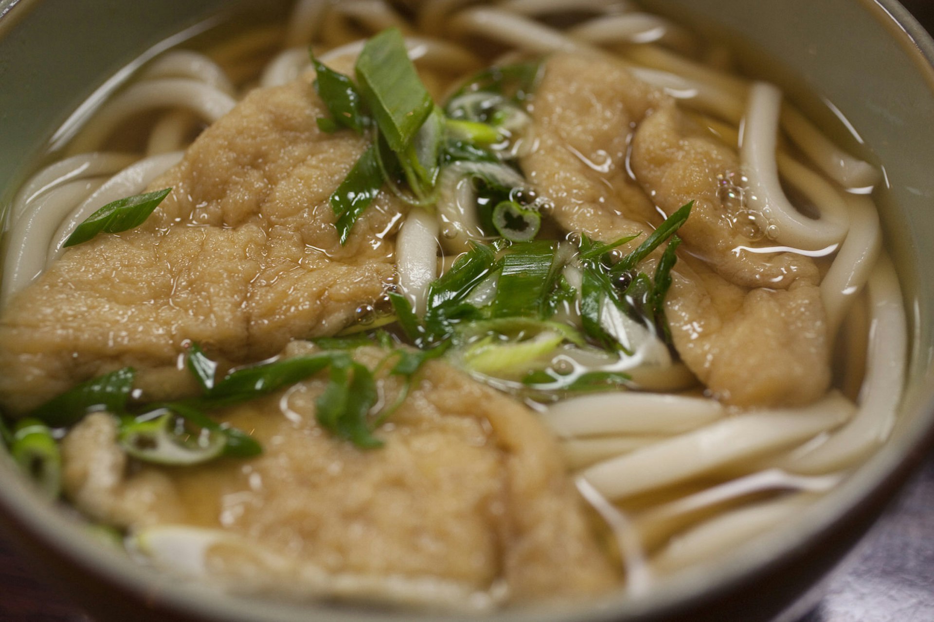 A close-up of a bowl of kitsune udon, showing tofu, noodles and dashi stock