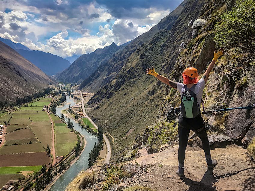 A girl with short orange hair stands on a high rock ledge with her back to camera, gazing out over a river in the Sacred Valley in Peru