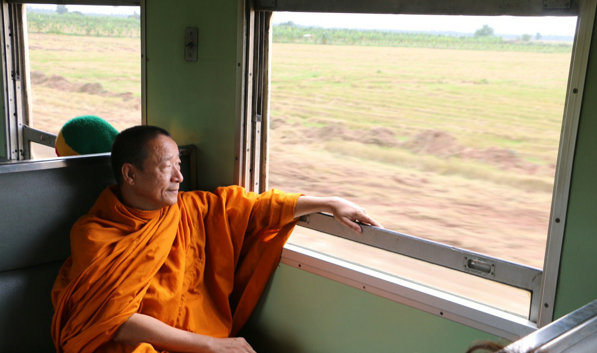 A monk looks out of the window on a train in Thailand rolling past green countryside