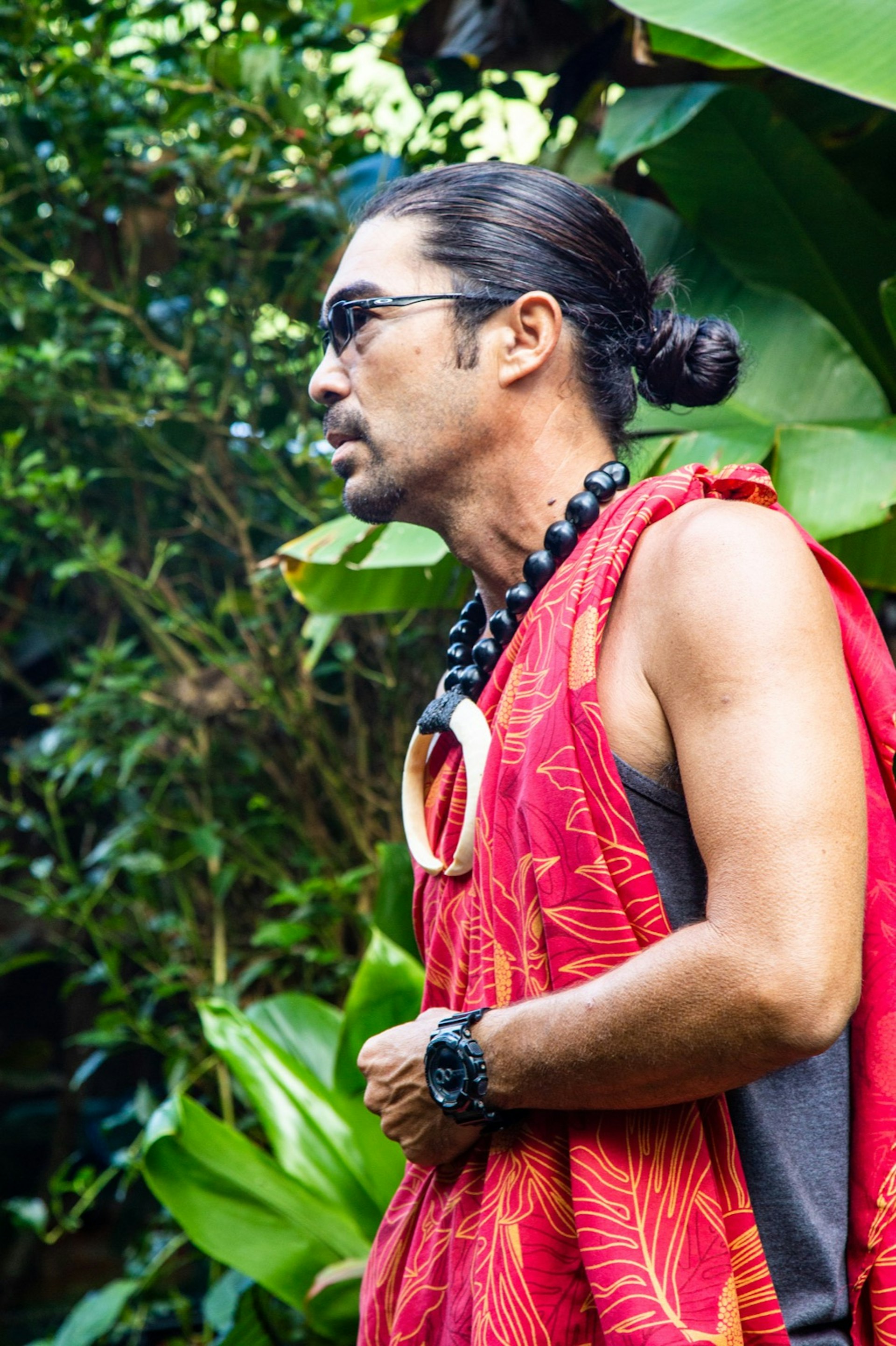 A man leads a group through a tropical jungle, dressed in a traditional Hawaiian tunic with a large necklace and his hair in a tight bun. He also wears modern sunglasses and a heavy modern watch