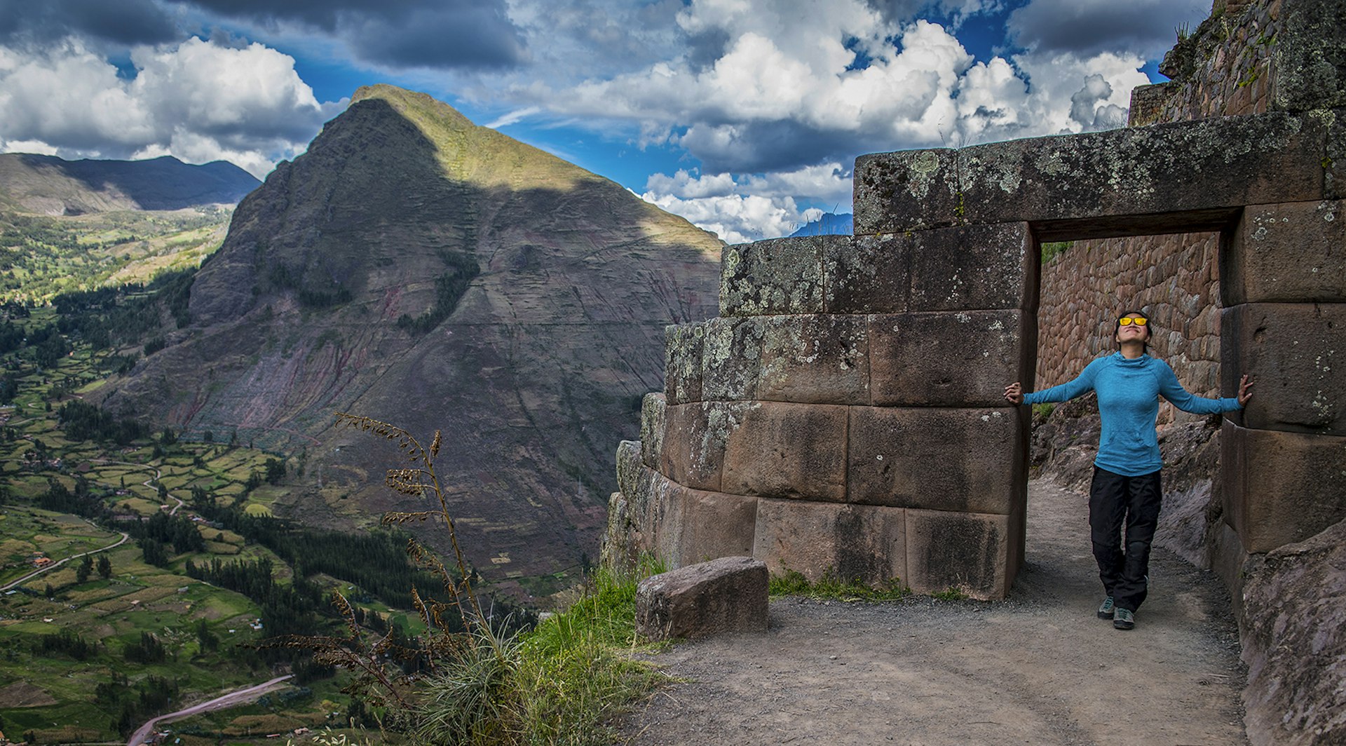 A woman stands in a stone doorway overlooking a green valley next to a rocky mountain