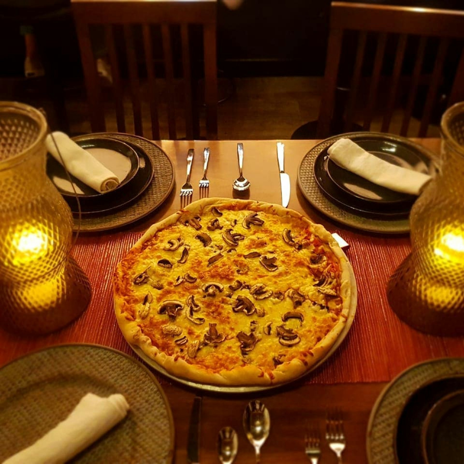 A pizza sits in the middle of a table at El Taller restaurant surrounded by plates and silver utensils. Everything is illuminated by a pair of candles in glass containers. 