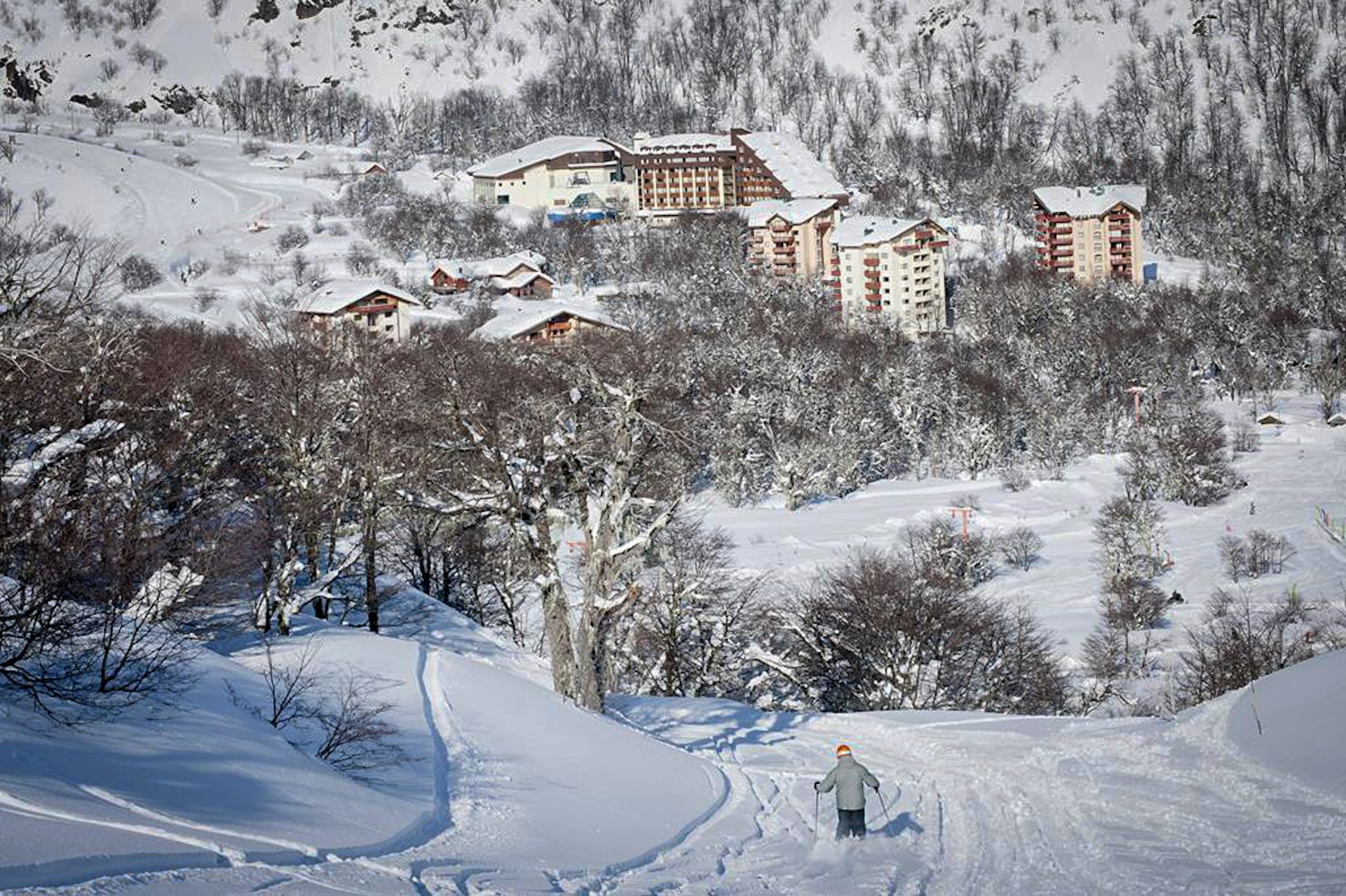 A skier skies down a hill towards a cluster of trees surrounding resort buildings at Termas de Chillán, Chile