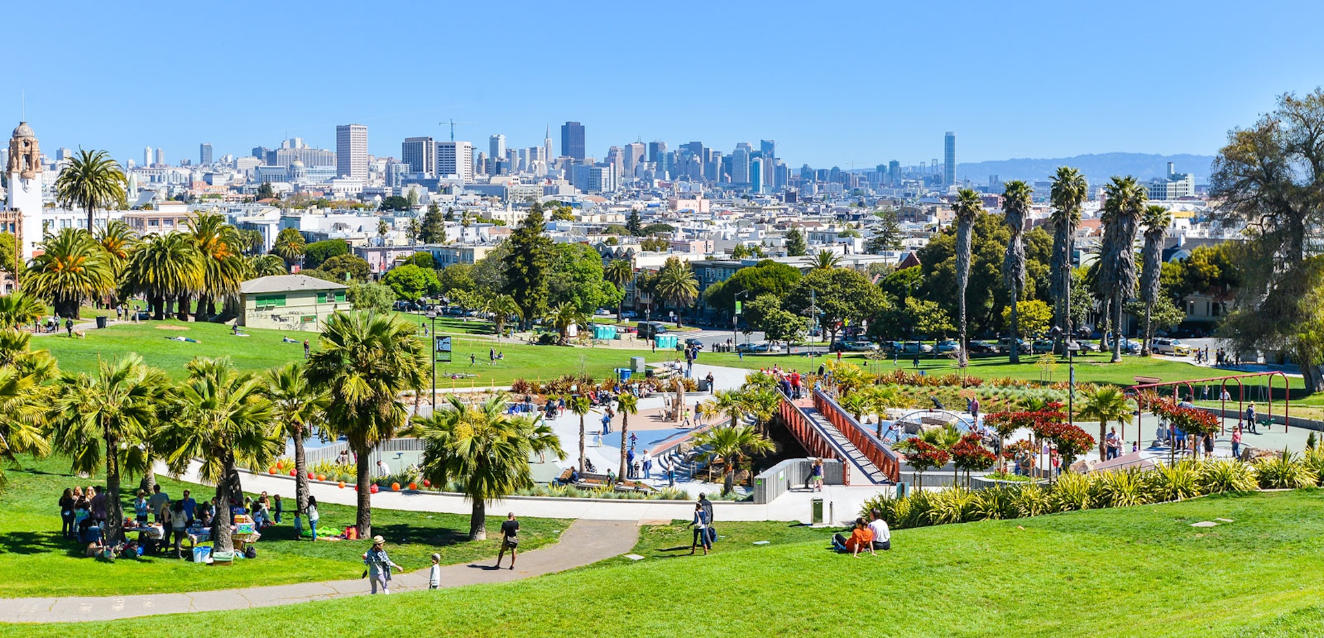 People play in a grassy park on a sunny day with the San Francisco skyline in the distance