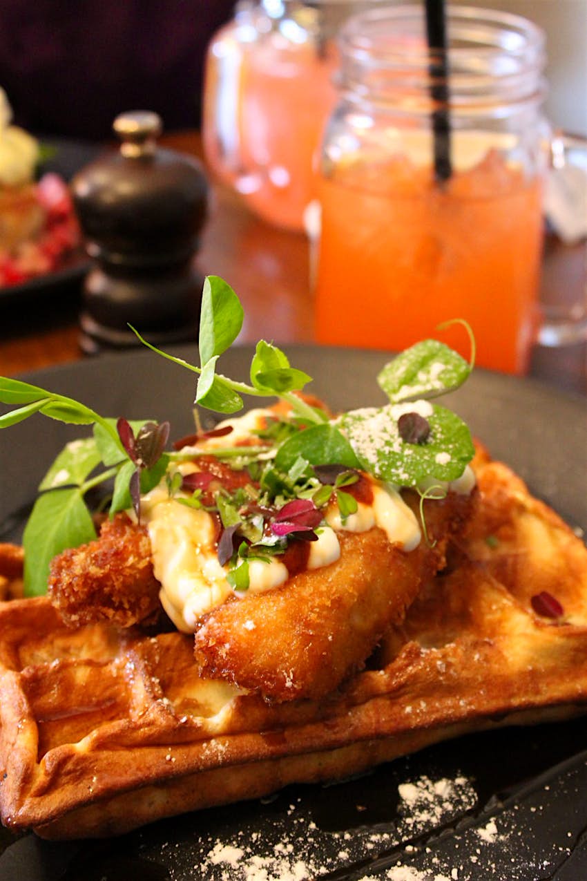 Close-up of Union Kitchen's brunch dish of fried chicken and waffle topped with a salad garnish, with drinks in mason jars in soft focus behind.