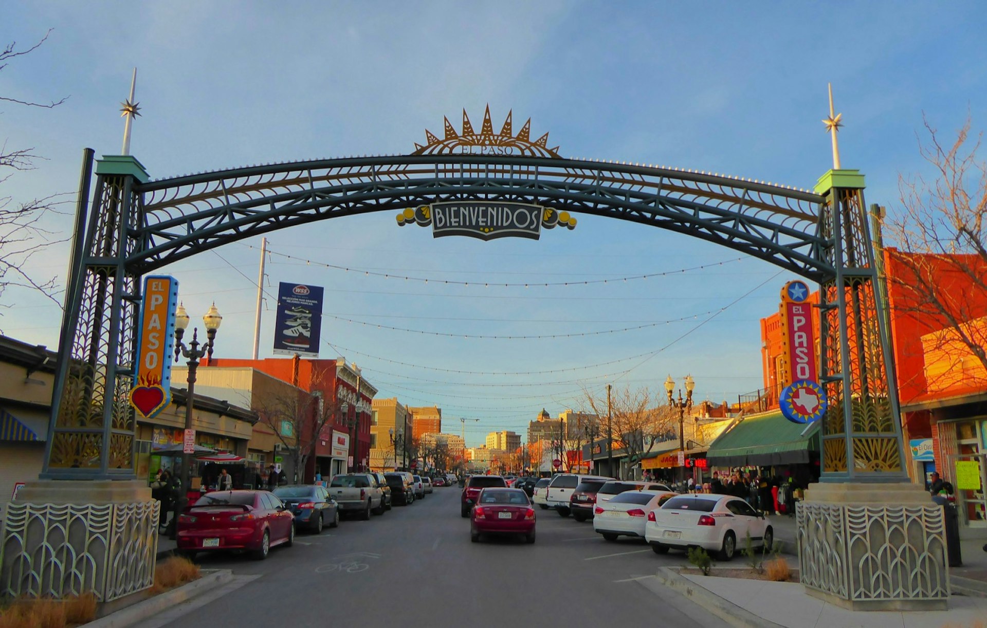 A city street, with shops along each side, is dominated by a colorful archway in the foreground that welcomes visitors to El Paso, Texas