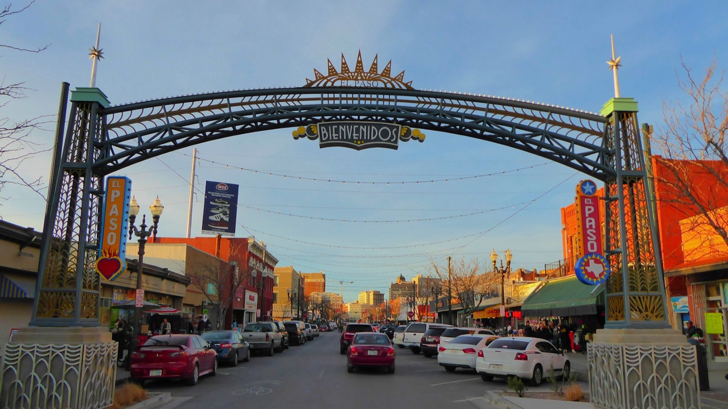 A city street, with shops along each side, is dominated by a colorful archway in the foreground that welcomes visitors to El Paso, Texas