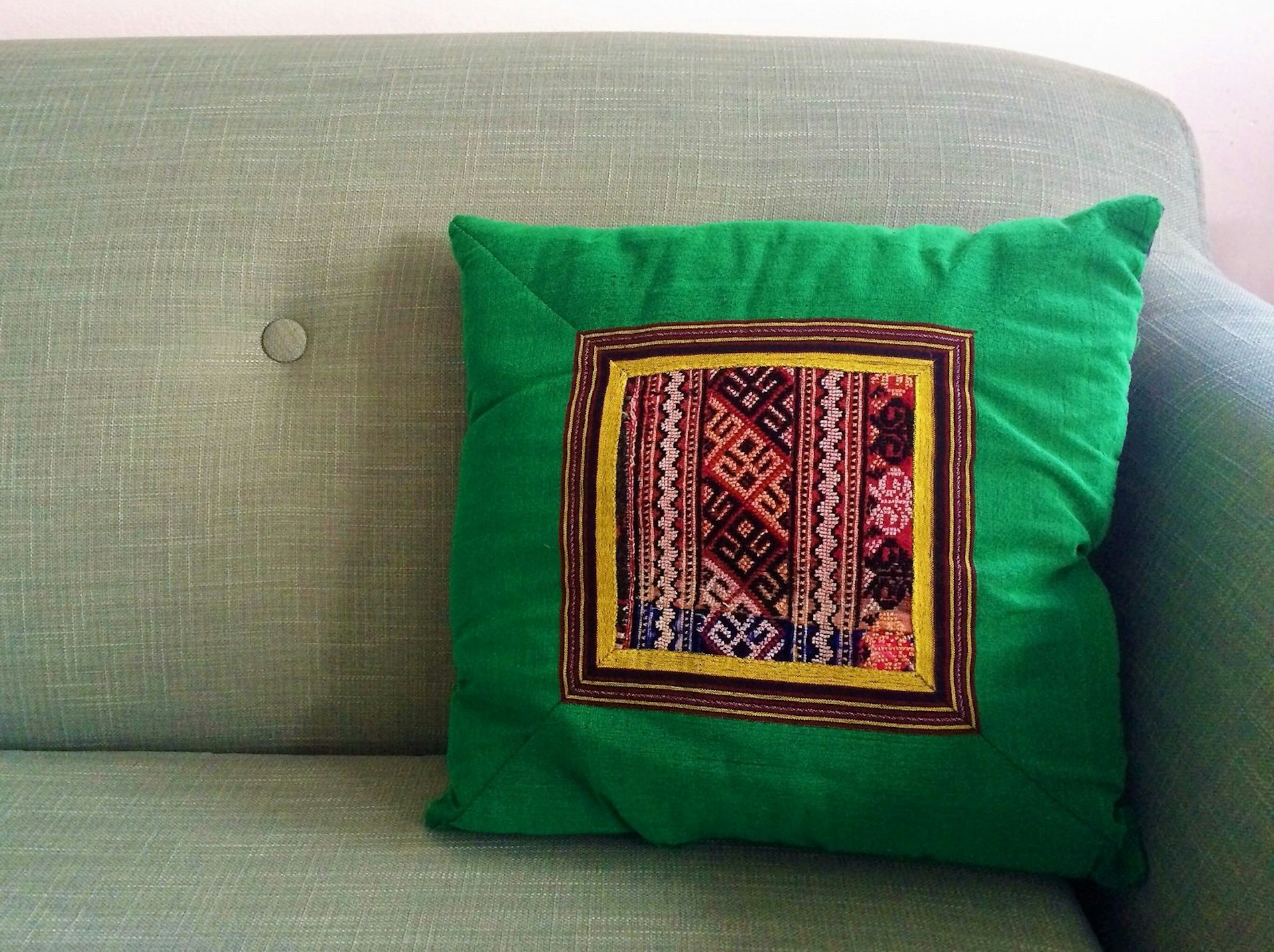 A green cushion with a patterned square in the middle resting on the arm of a sofa