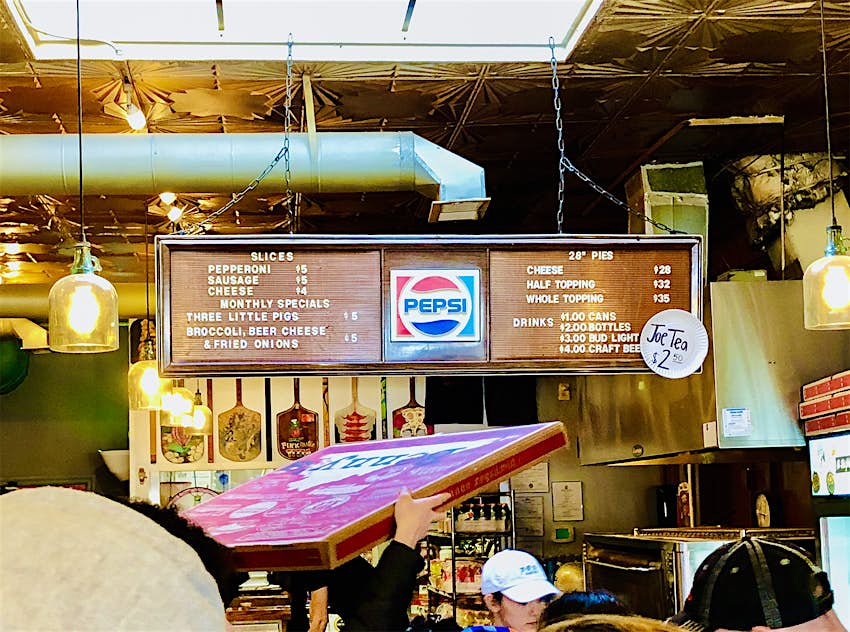 Interior shot of a high-ceilinged building with tin ceiling tiles and a crowd of people looking at a menu board with pizza options and prices