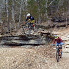 A mountain biker leaps off a rocky outcropping while a second rider takes a side option of a curving rock path