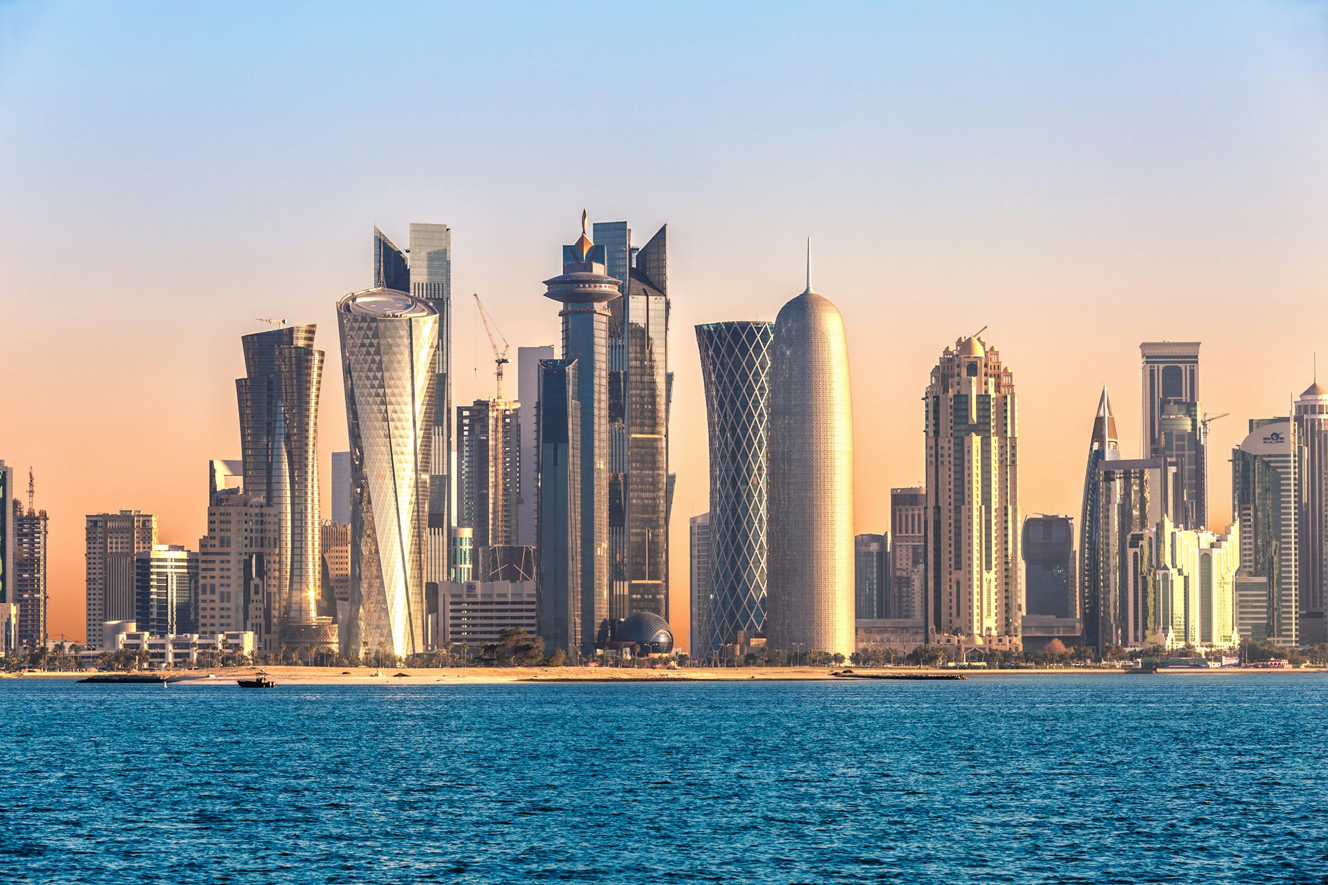 The skyline of Doha, Qatar, as seen from the water at sunset; numerous skyscrapers have unique, twisting shapes.