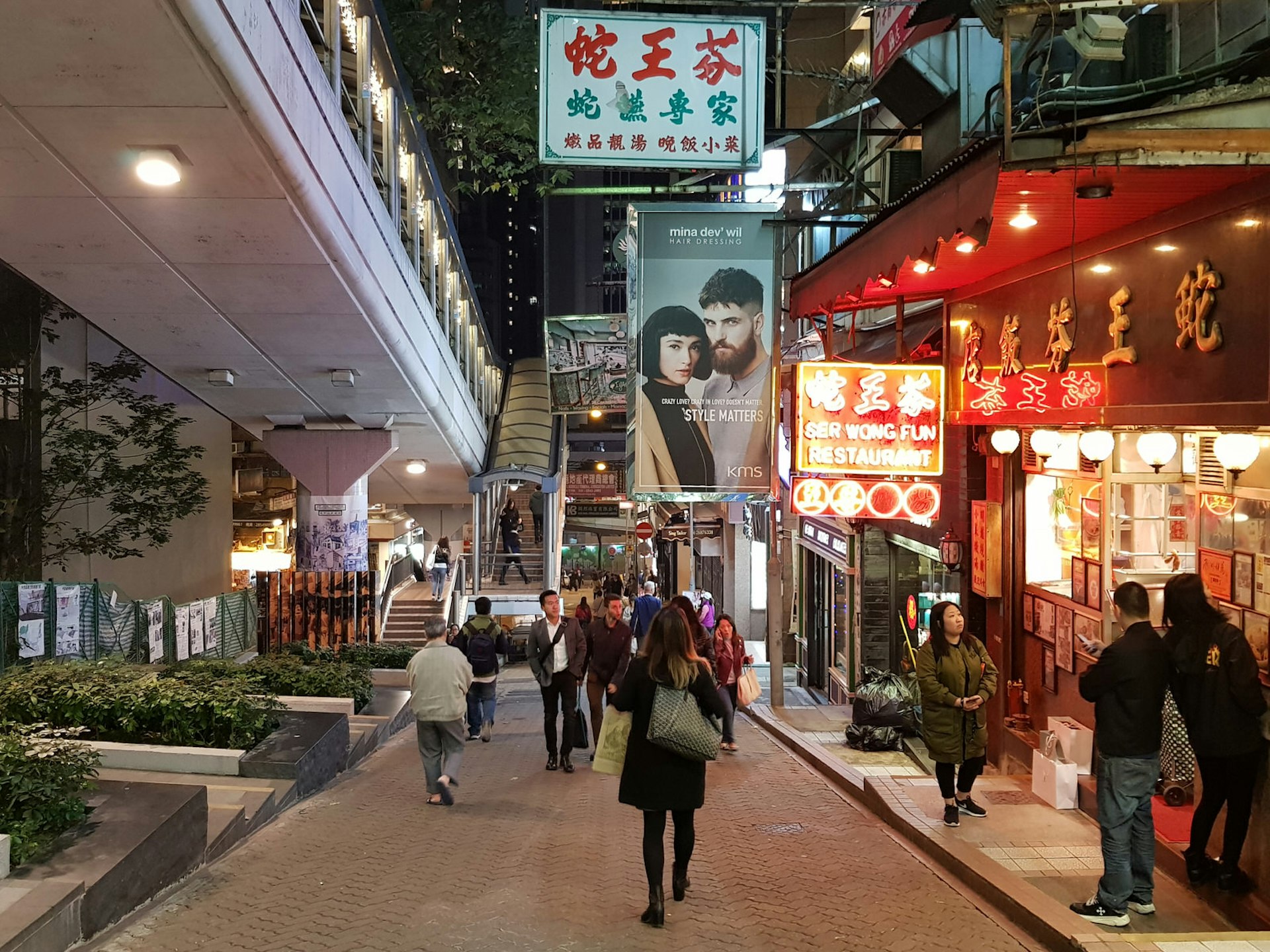 People walk down a pedestrian street lit on one side by traditional shops, with the escalator overpass above to the left.