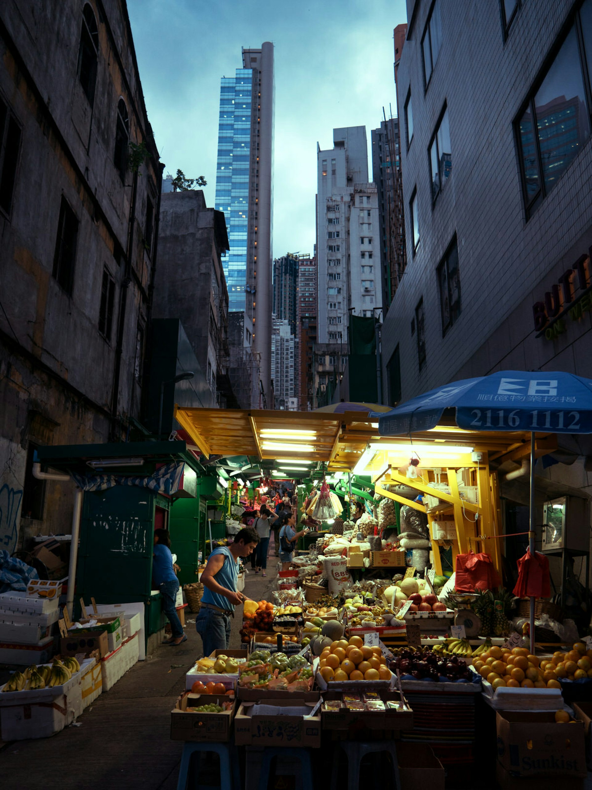 Small stalls with awnings and bare bulbs sell fruit and veg on Graham Street in the shadow of high-rise buildings