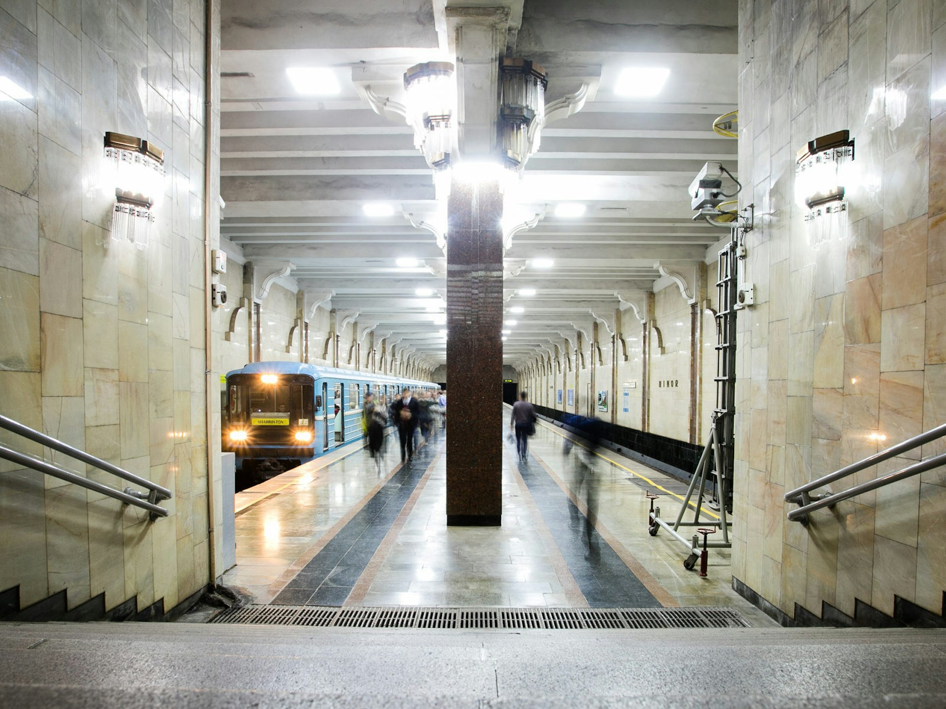 View of Minor station on the Tashkent Metro with a blue train passing through the platform on the left and bright white lights illuminating the floor.