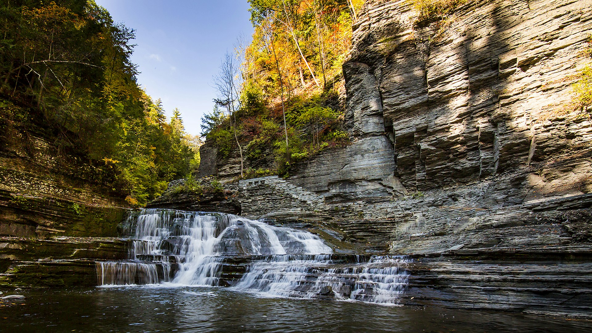 Waterfall cascading through a rugged stone gorge surrounded by fall foliage on a sunny fall day