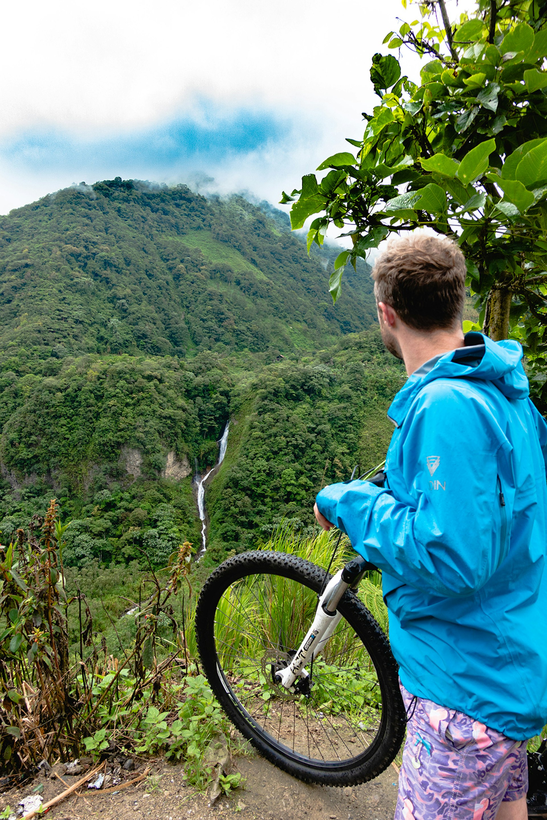 A man holds a bicycle and looks out over a green hill with a waterfall running in the middle.
