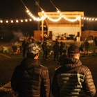 Two men in outdoors gear walk toward a small stage at night, as strings of globe lamps light their way