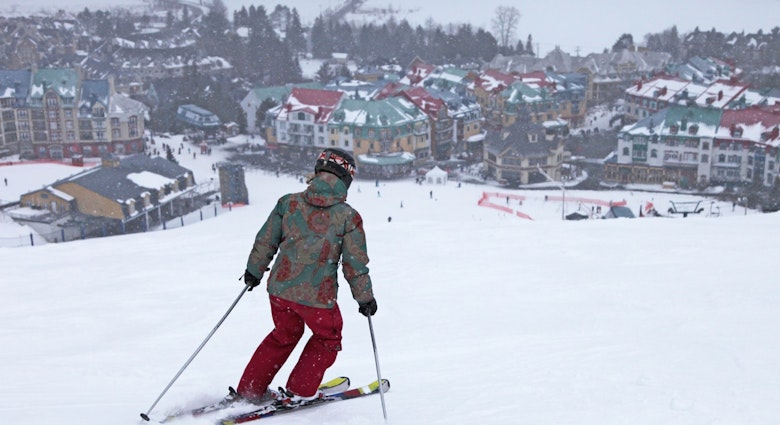 A woman skis on top of a mountain as a picturesque village, covered in snow, spreads out below her at the foot of the slope