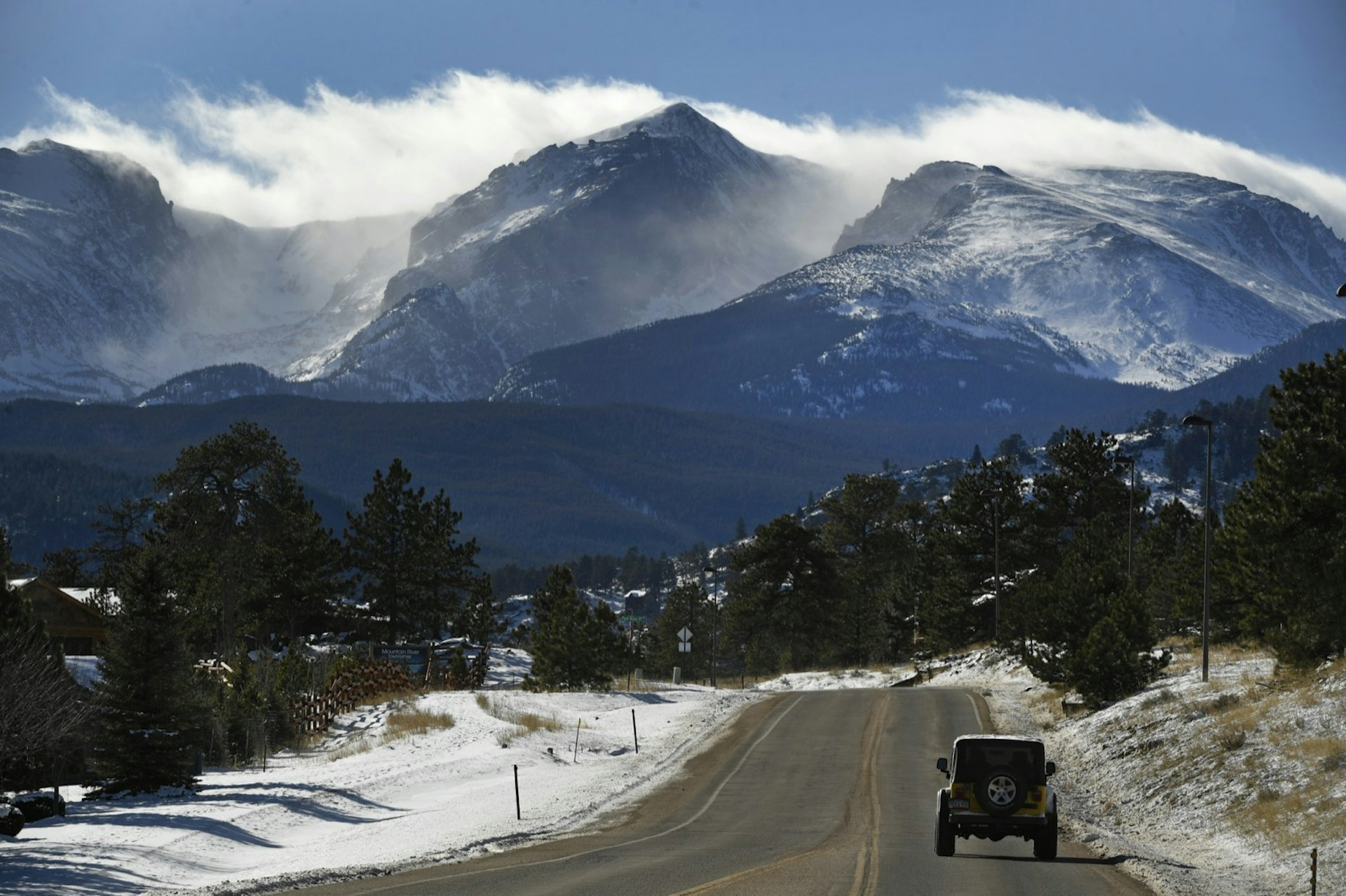 A jeep wrangler drives down a road with snow on both sides, as the rocky mountains loom ahead and snow is blown off the tops of the peaks