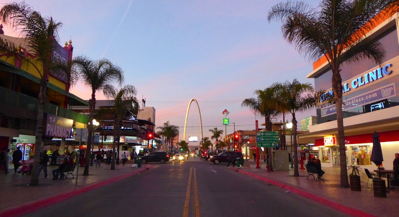 An empty street lined with palm trees with an arch at one end under a blue sky dotted with pink clouds
