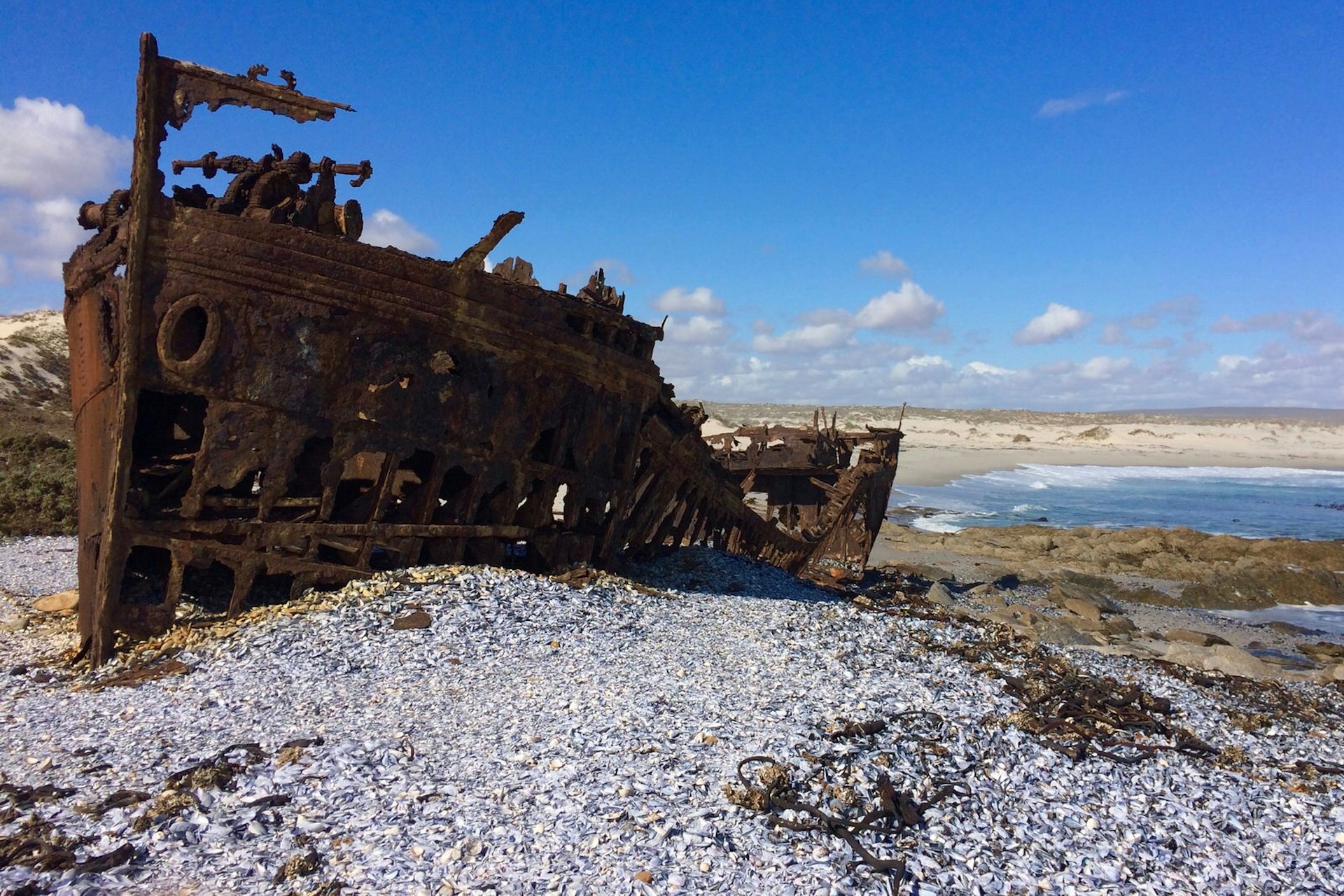 A huge rusty shipwreck sits on a bed of shells, with a beach in the distance