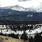 Two deer graze in the foreground as snowy peaks surround a brown valley in Estes Park Colorado