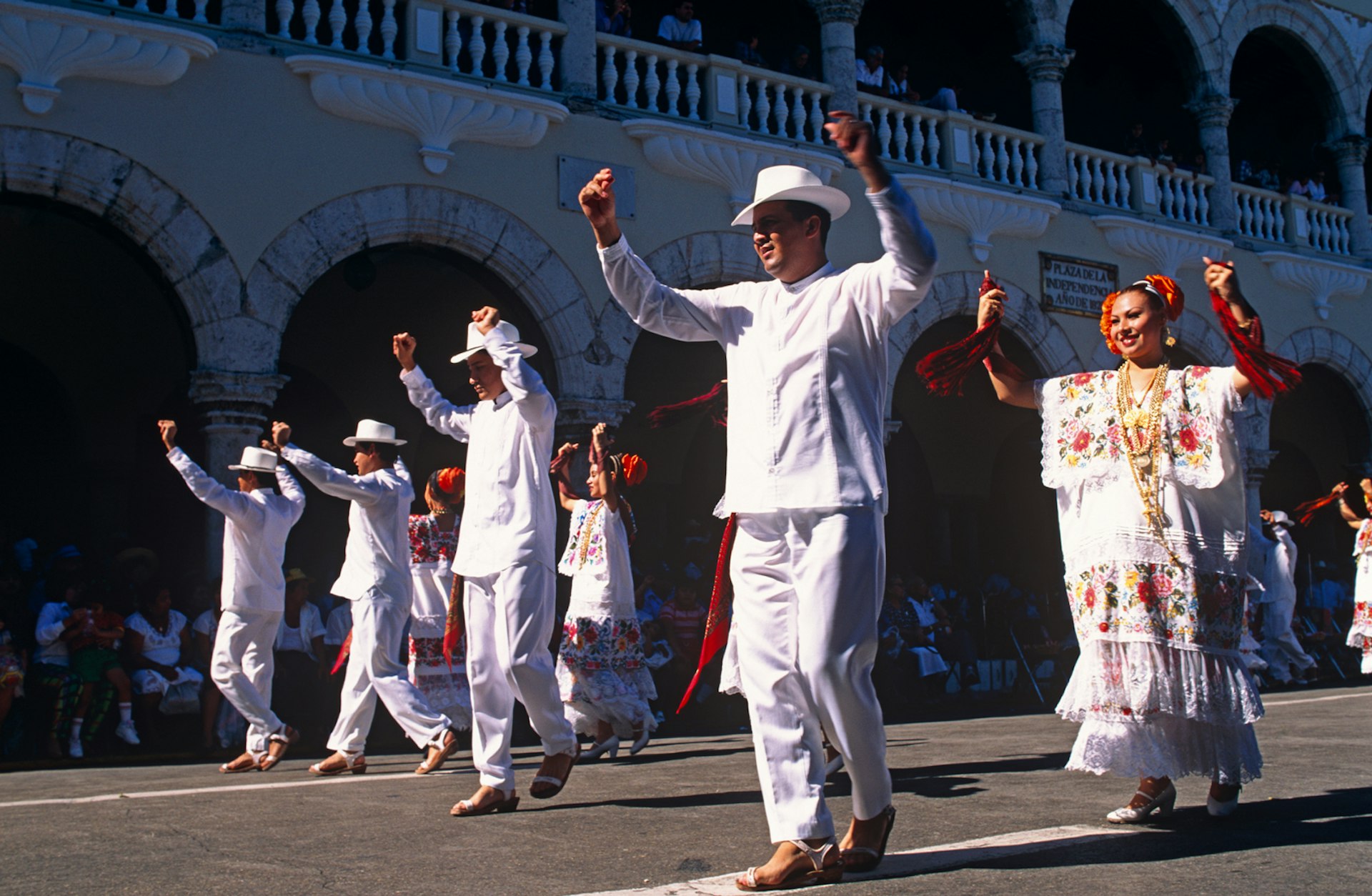 Men in white outfits and women in colorfully embroidered dresses perform in the street in front of a colonial building