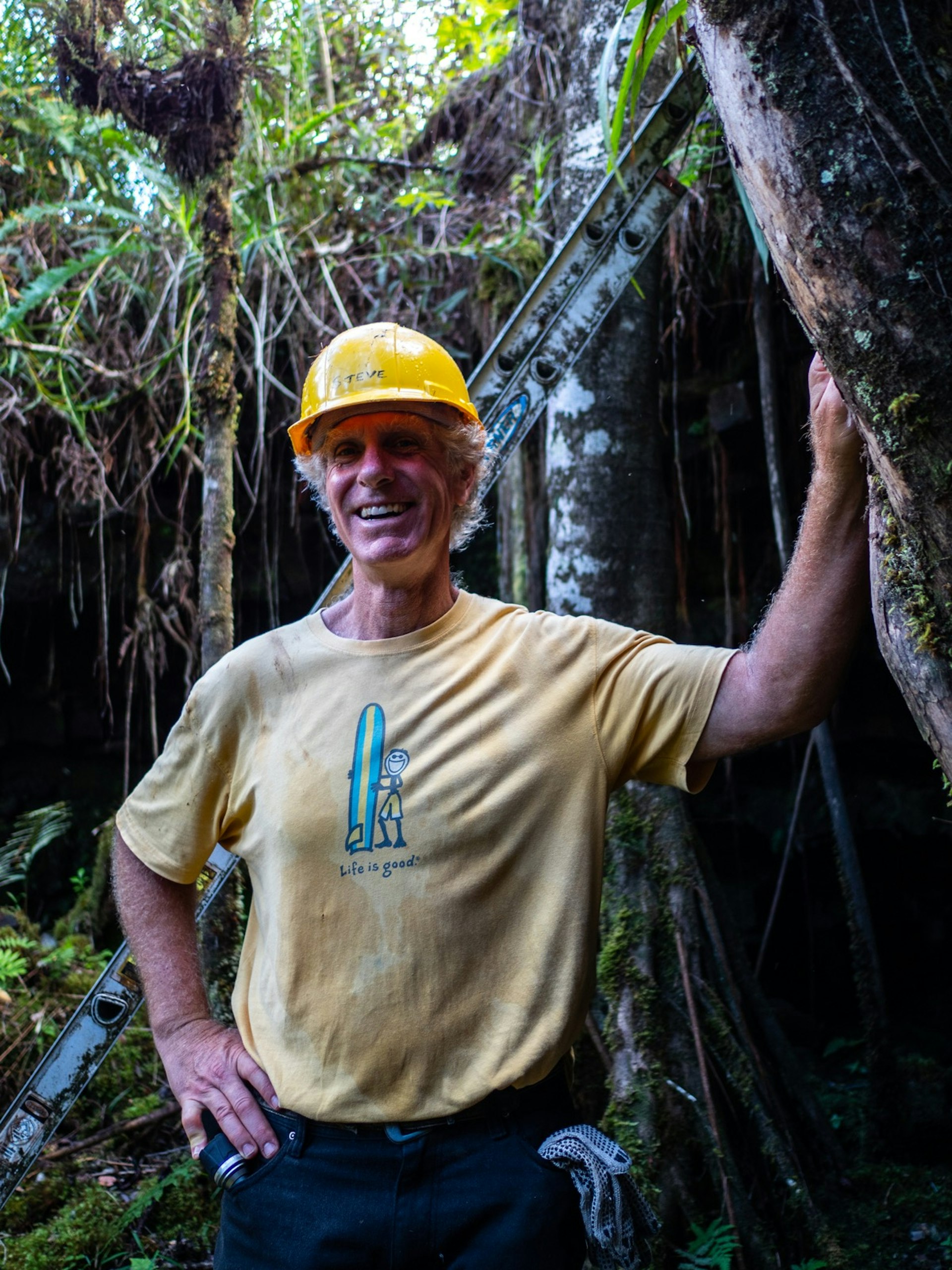 A smiling man with a yellow hard hat and a shirt that says life is good stands outside a cave entrance in the jungle