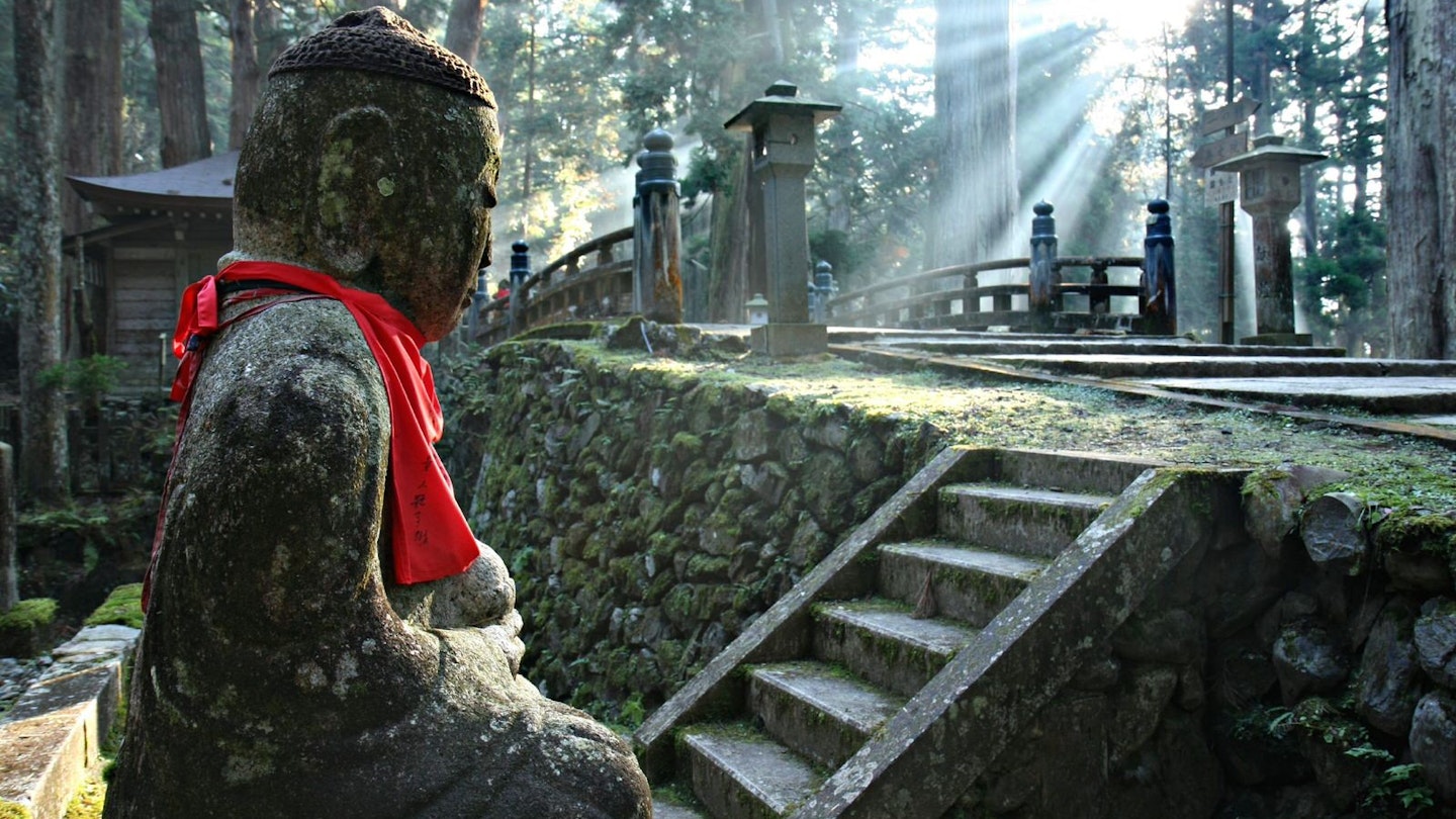 A statue stands in Oku-no-in as dappled sunlight shines through the trees