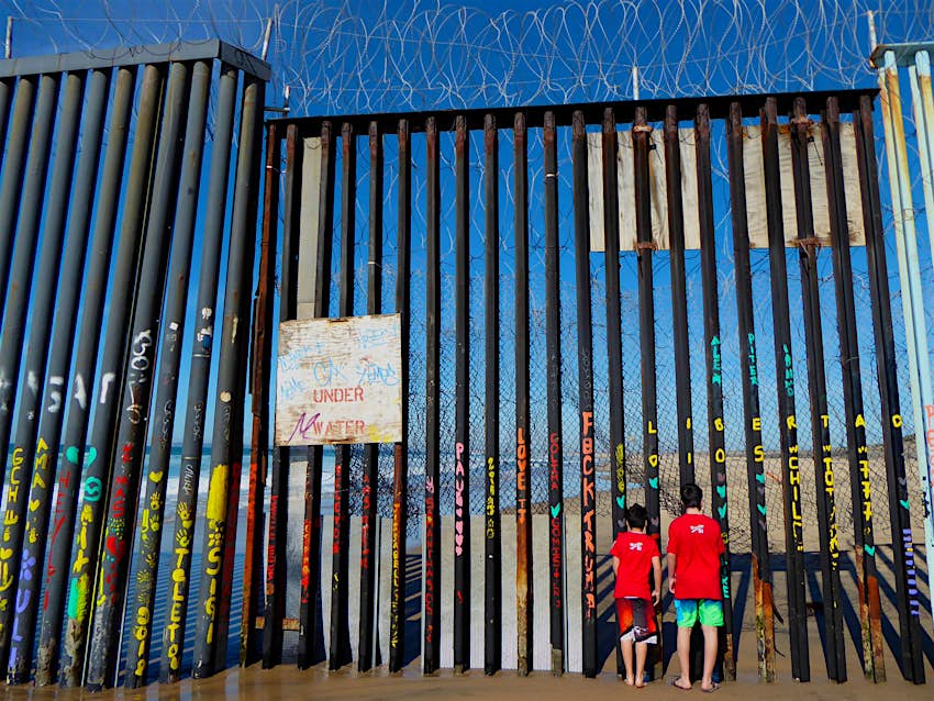 Two boys in red t-shirts stare through a wall of steel bars heavily covered in graffiti with chicken wire on the top