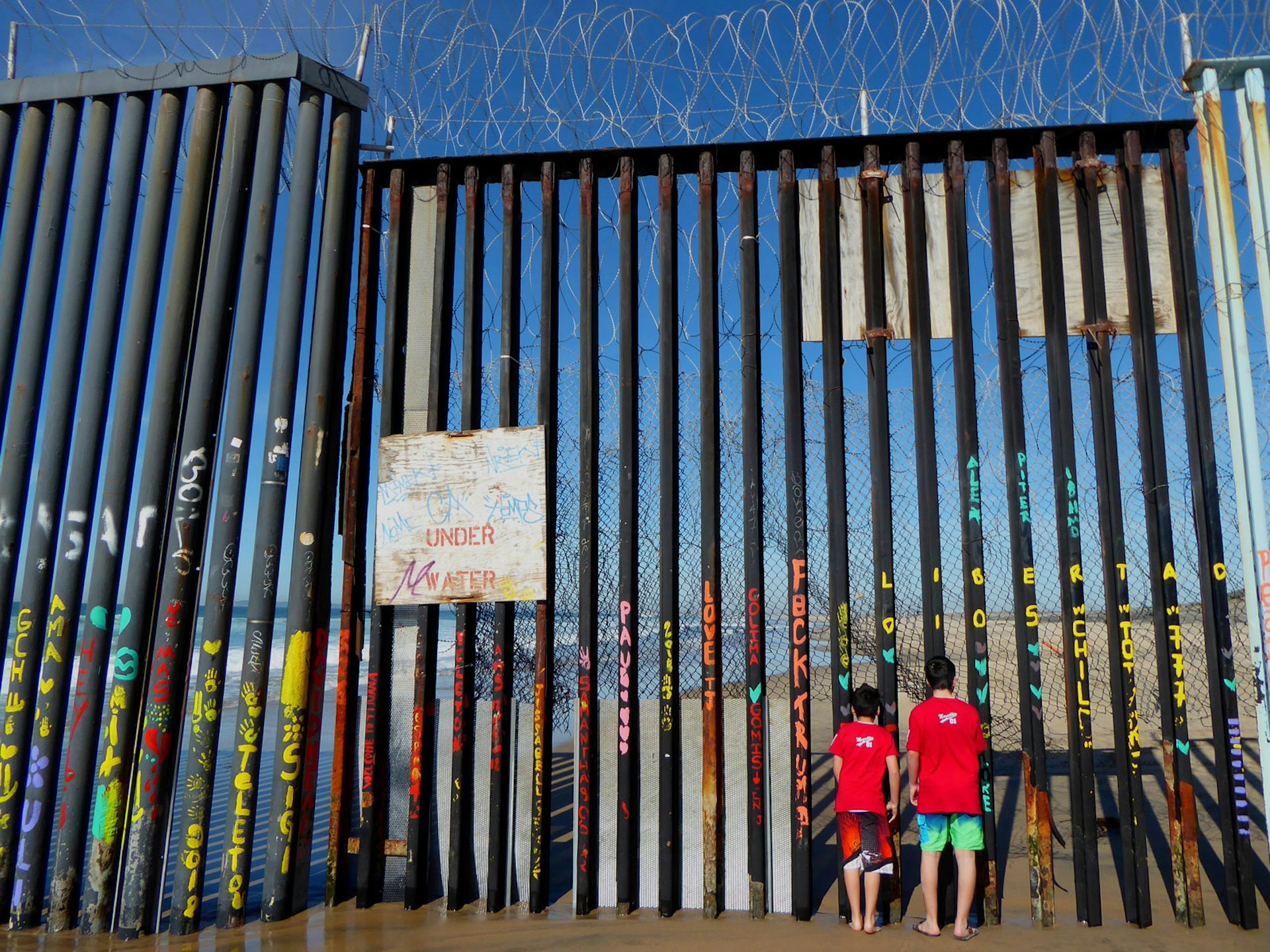 Two boys in red t-shirts stare through a wall of steel bars heavily covered in graffiti with chicken wire on the top