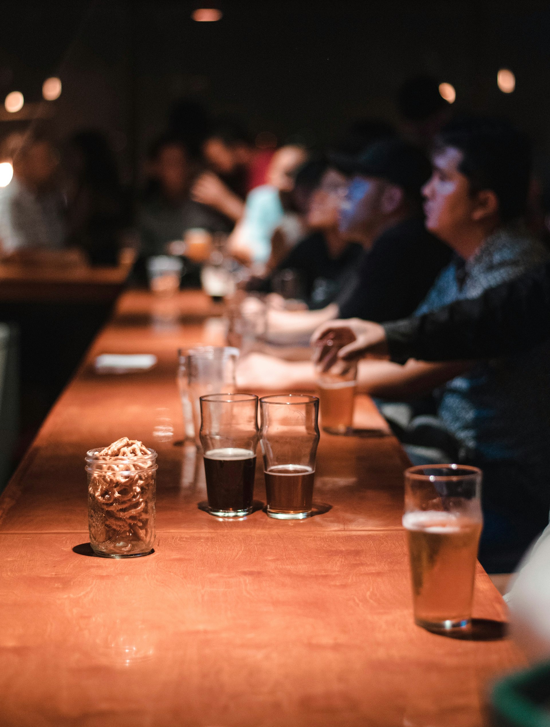 Half full glasses of beer in multiple hues sit on a bar lined with sitting men