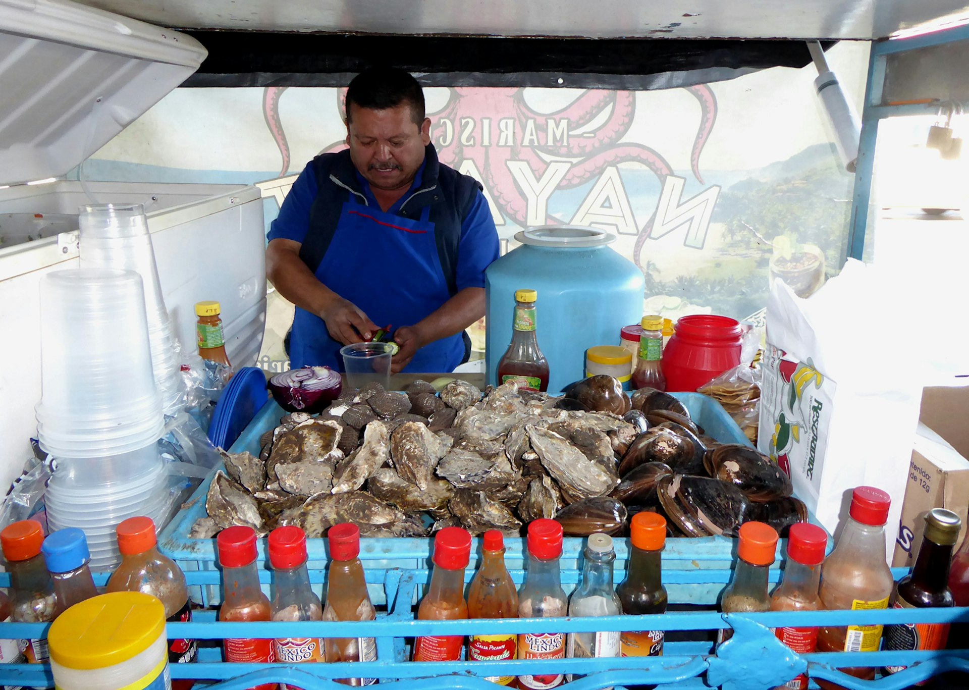 A man shucks oysters behind a giant pile of oysters and a plethora of sauces