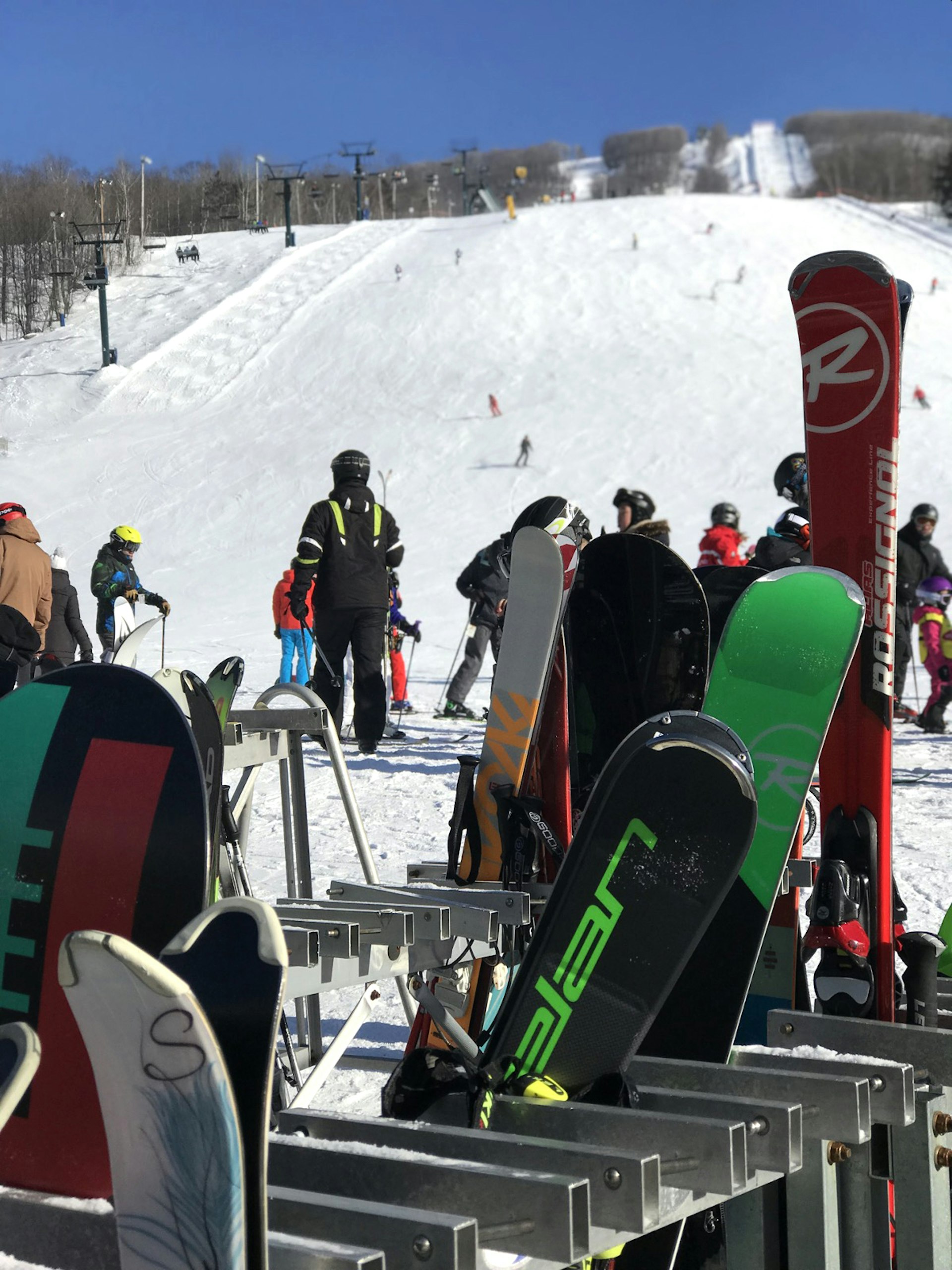 A rack of skis and snowboards are in the foreground and snow sports enthusiasts are in the background at the foot of a pristine, groomed ski slope in Mont-Tremblant, Quebec.
