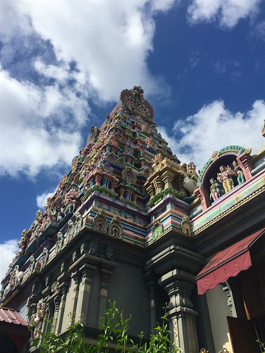 Under a blue sky dotted with white, cotton ball-looking clouds, is the tapering tower of the Hindu Sri Navasakthi Vinyagar Temple. The four-sided pyramid is adorned with numerous layers of colourful engravings © Matt Phillips / Lonely Planet