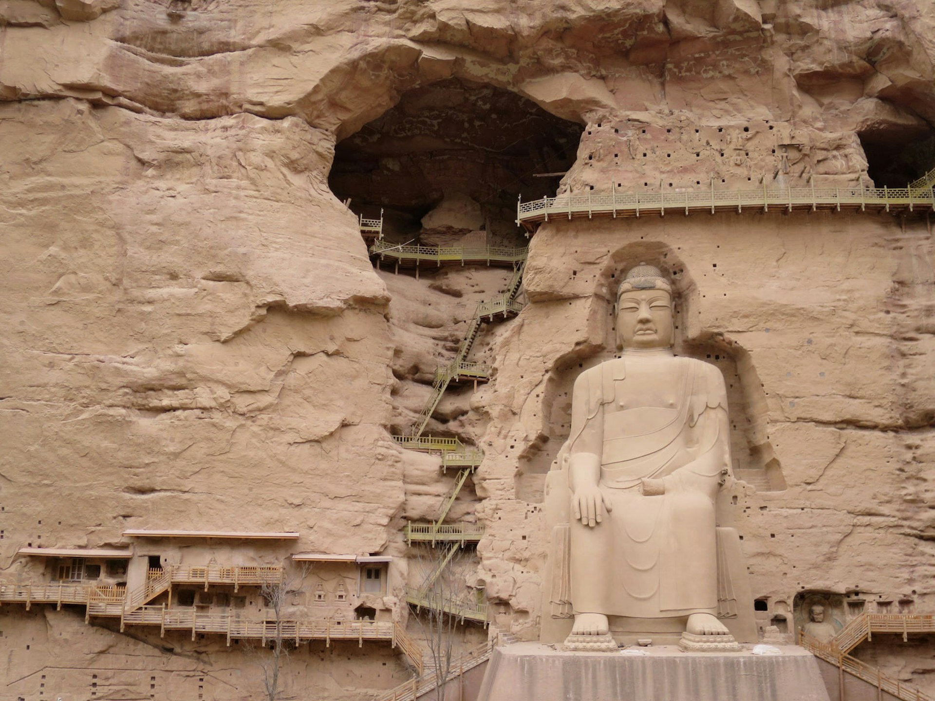 A huge seated Buddha statue carved into a sheer desert cliff; to the left, a high-up cave connected by a series of ladders and walkways.