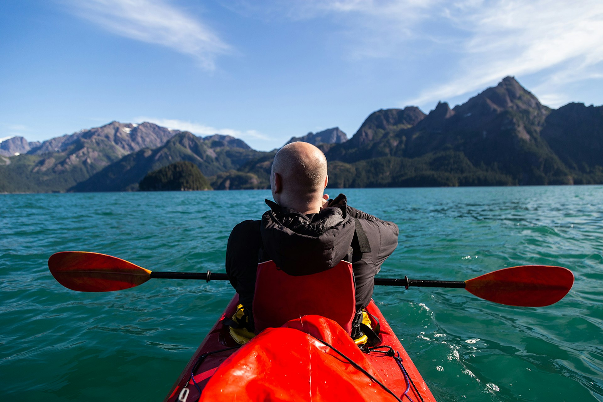 A kayaker in a red kayak looks across turquoise water to rugged mountain peaks