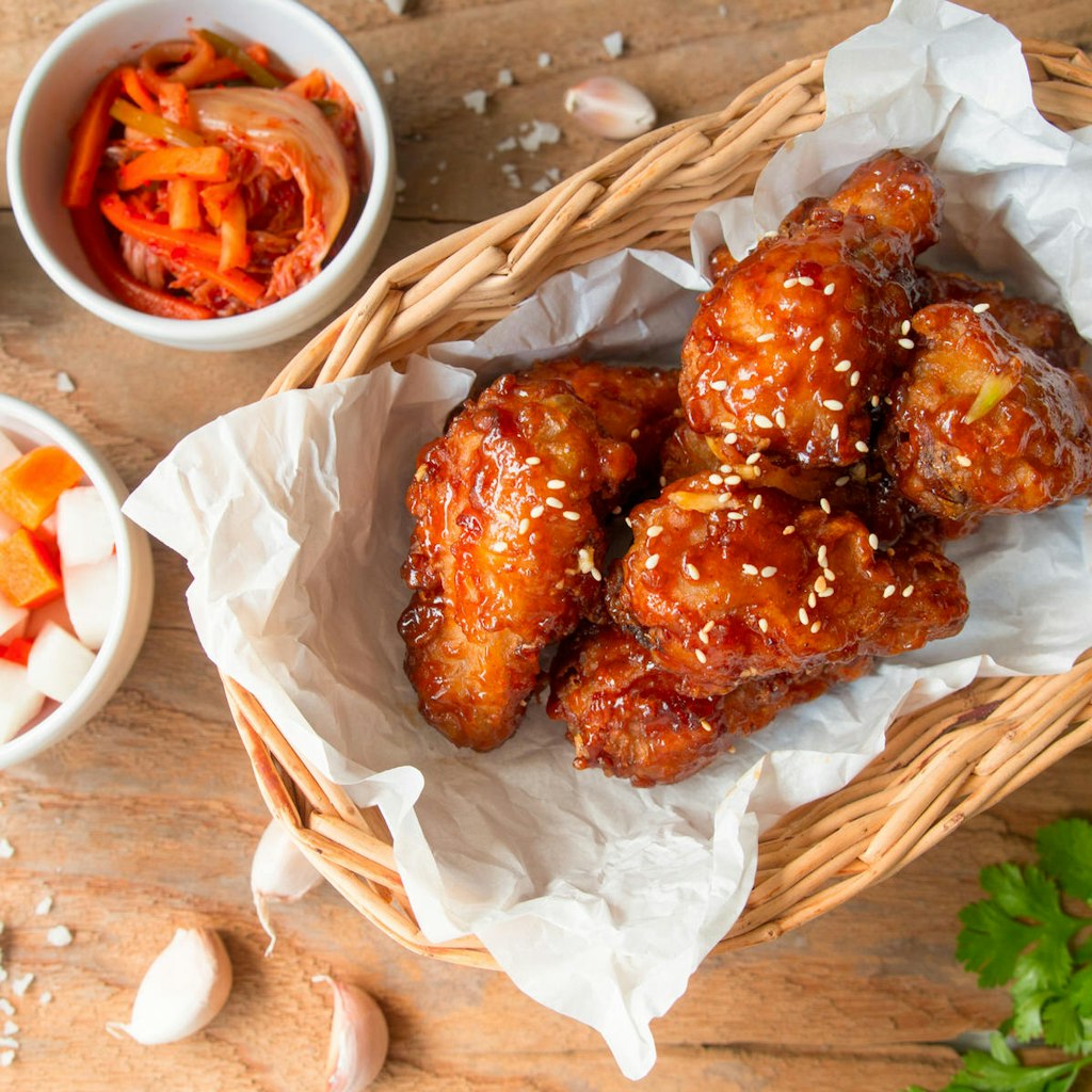 A basket of Korean fried chicken with sides of pickled radish and cloves of garlic on a wooden table