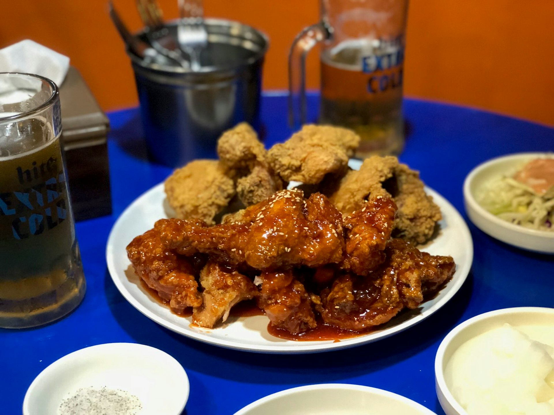 A plate of Korean fried chicken covered with red sauce on a blue table with mugs of beer in the background