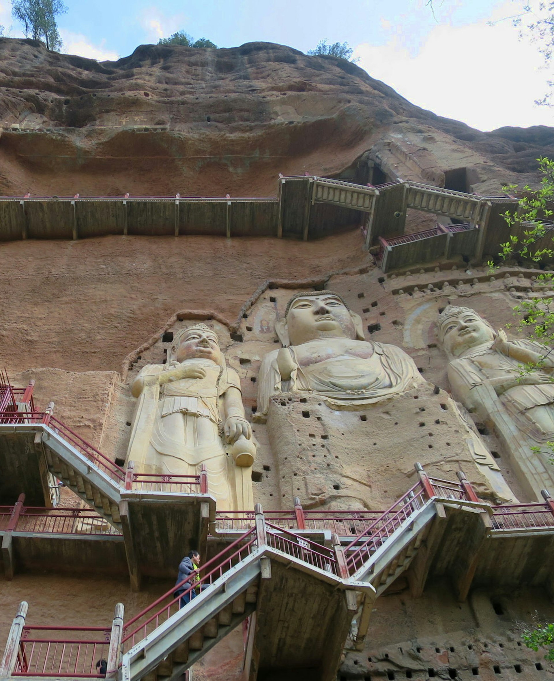 A tall Buddha carved into a red cliffside flanked by two bodhisattvas and surrounded by steep stairways.